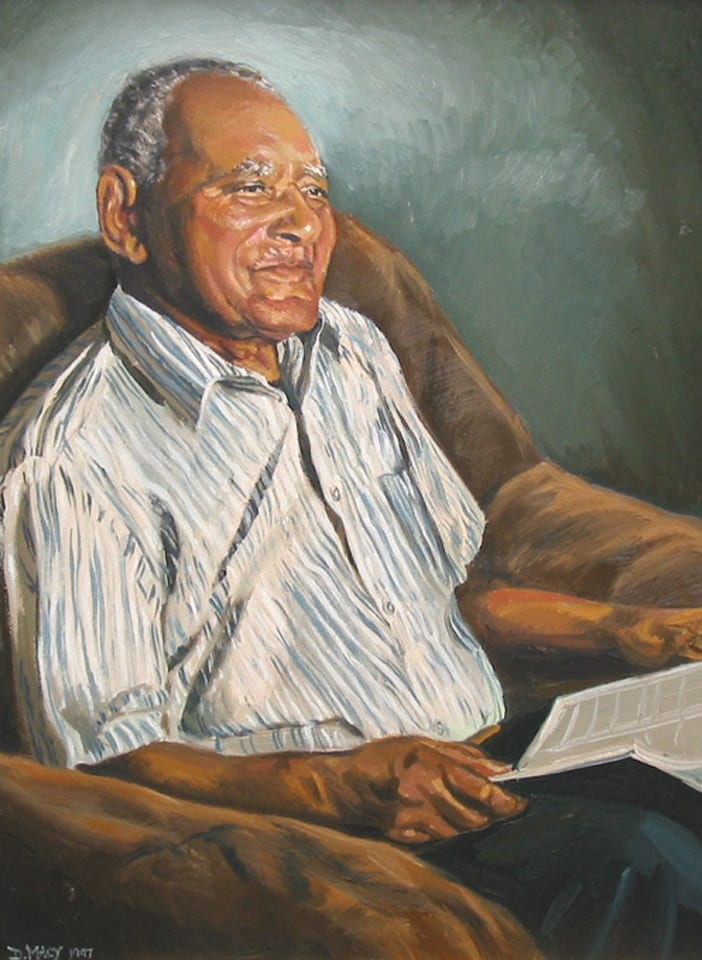 This portrait of the last living New Bedford whaleman Antonio L. Lopes shows him at the age of 100. He is an elderly man, smiling, with receding gray hair, and wearing a striped short-sleeved shirt. He is seated in a brown overstuffed chair with a book in his lap.