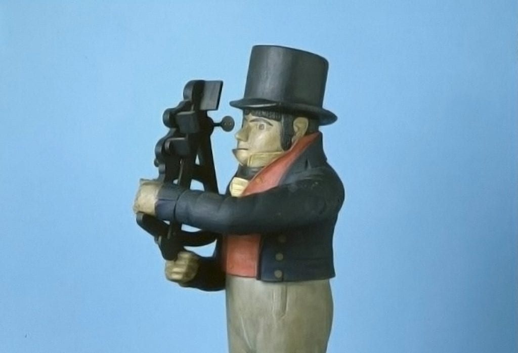 A full figure wooden navigator holding black quadrant, sight at right eye, right hand on index arm, frame in left hand. The Little Navigator is wearing a black short jacket with yellow buttons; a red waistcoat with yellow buttons; a white shirt collar with a ruffle; a black band under the collar; a black stovepipe hat; grey pants; and black boots. He has black square-cut banks, long sideburns, arching eyebrows, thin lips, and a small pointed nose. He is standing on a square black box.