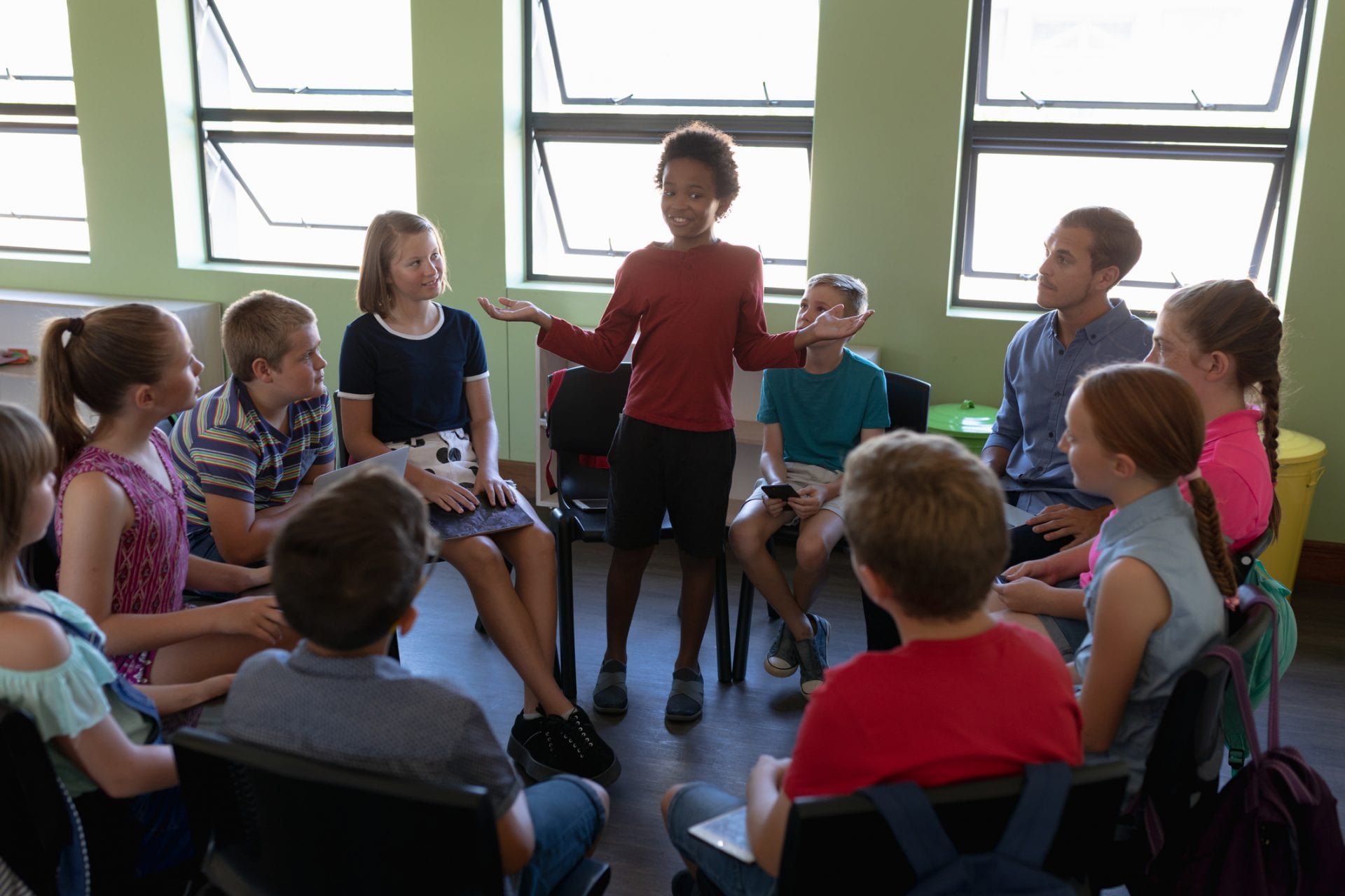 Front view of a diverse group of elementary school kids sitting on chairs in a circle and interacting during a lesson, one African American girl standing and talking while her classmates and male Caucasian teacher sit and listen