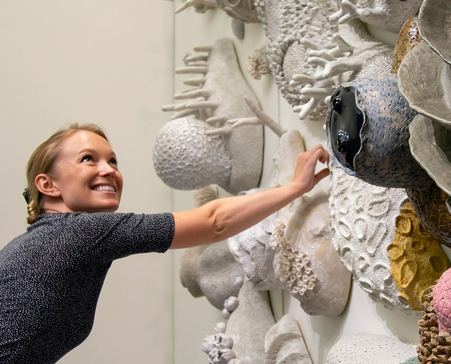 A photograph of a woman reaching out to touch a wall sculpture of coral.