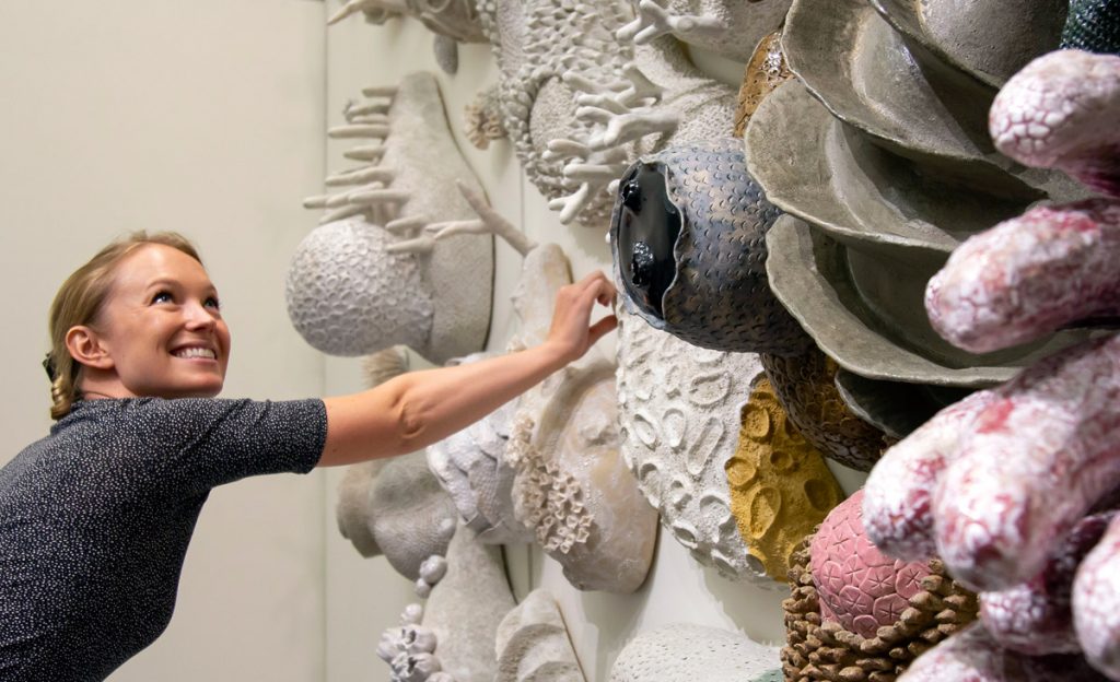 A photograph of a woman reaching out to touch a wall sculpture of coral.