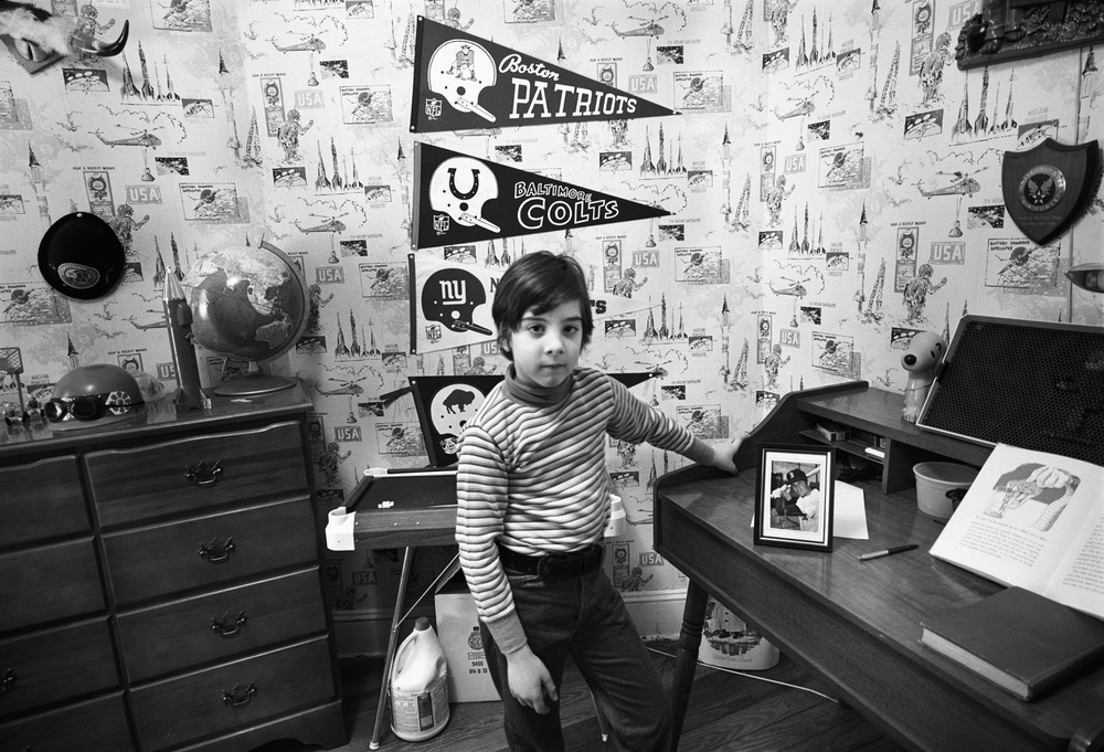 A black and white photograph; a young boy poses for a picture in his room. Behind him is a cartoonish wallpaper and sports memorabilia.