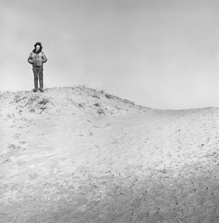 A black and white far shot of a man standing on a sandy hill at a beach.