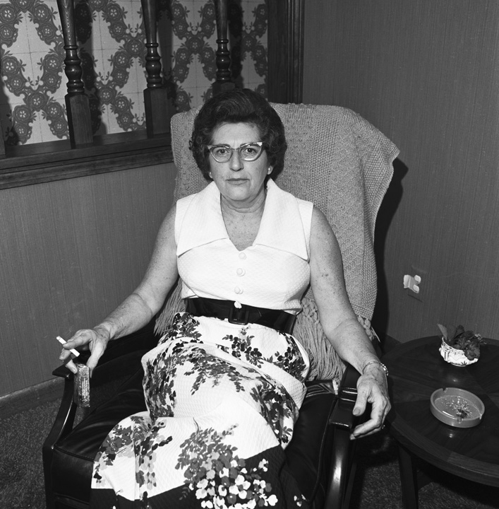 A black and white wide shot of a woman sitting in an armchair posing for the camera. She is holding a cigarette in her hand.