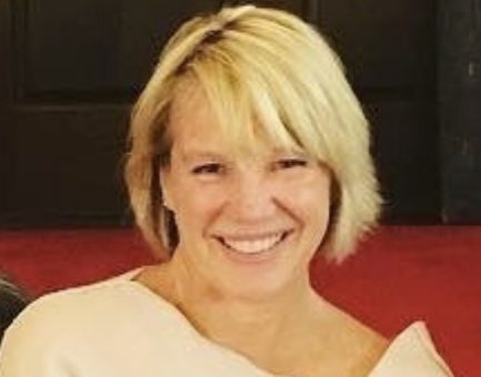 A bust shot of an older woman with short blonde hair and a beige dress. She is looking at the camera and smiling.