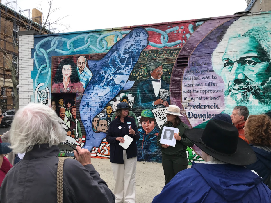 A group on the Lighting the Way to Justice walking tour. The group stands listening to the two guides in front of a social justice mural downtown.