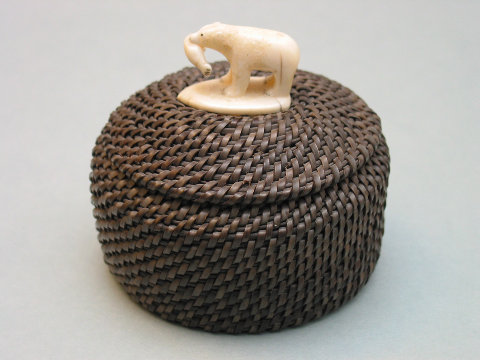 A basket woven using whale baleen, with a decorative polar bear at the top with a fish in it's mouth, made of ivory.