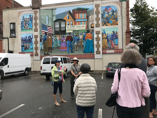 A group on the Women of the Underground Walking Tour. They are standing in a parking lot in front of the 54th regiment mural downtown. They are listening to the two tour guides, one of whom is a park ranger.