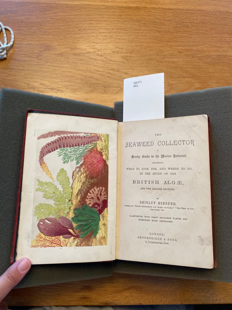 A book opened to its first page; on the left there is an illustration of different kinds of seaweed and algae, on the right reads the title "The seaweed collector, a handy guide to the marine botanist. Suggesting what to look for, and where to go, in the study of the British Algæ, and the British sponges" by Shirley Hibberd