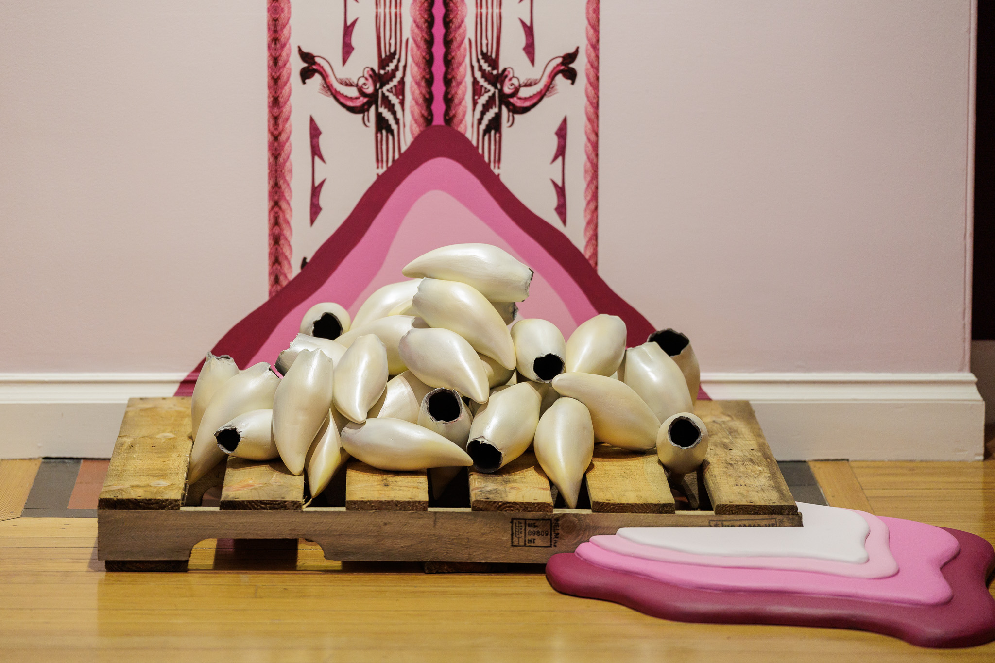 A collection of whale teeth sculptures arranged on a wooden pallet. The sculptures are displayed against a pink wall with decorative patterns. The background includes layered pink and white shapes.