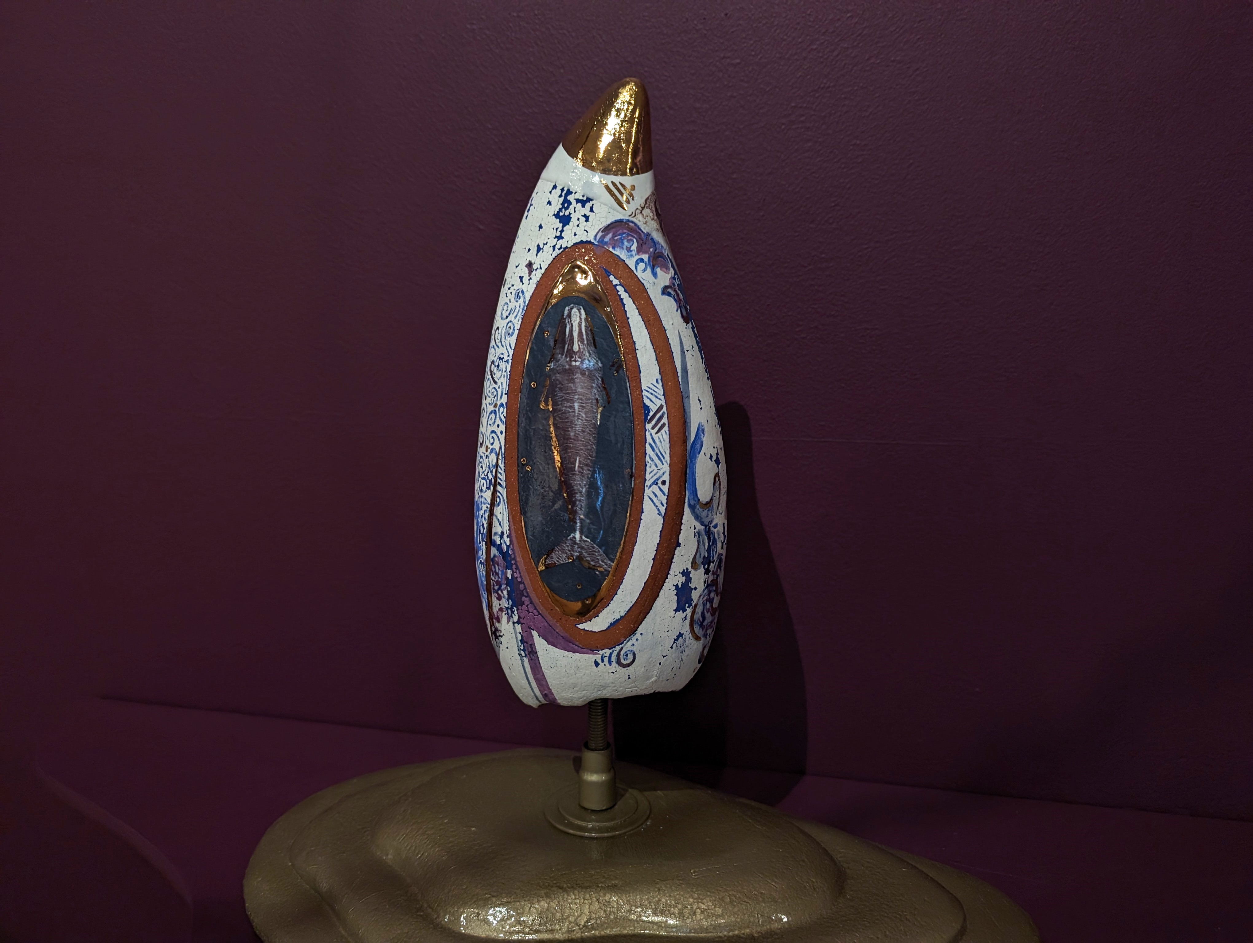 A sculptural whale tooth displayed against a dark purple wall. The object has an elongated, oval shape with a gold-painted top and intricate blue and white patterns on its surface. In the center, there is an oval illustration of a whale, framed with gold and brown. The object is mounted on a stand, showcasing its detailed and artistic design.