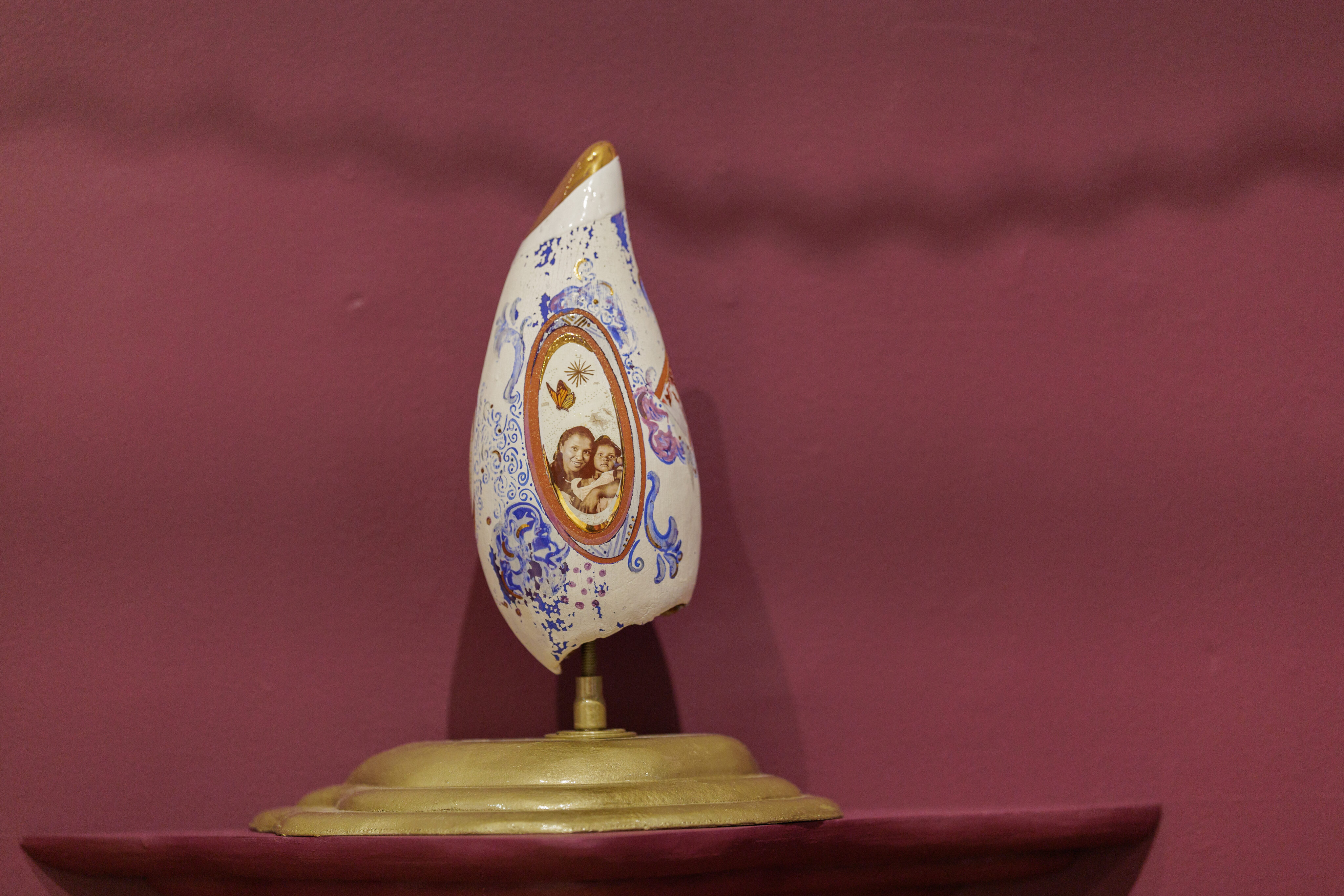 A sculptural whale tooth displayed against a pink wall. The object has an elongated, oval shape with a gold-painted top and intricate blue and white patterns on its surface. In the center, there is an oval illustration of the artist and her daughter smiling, framed with gold and brown. The object is mounted on a stand, showcasing its detailed and artistic design.