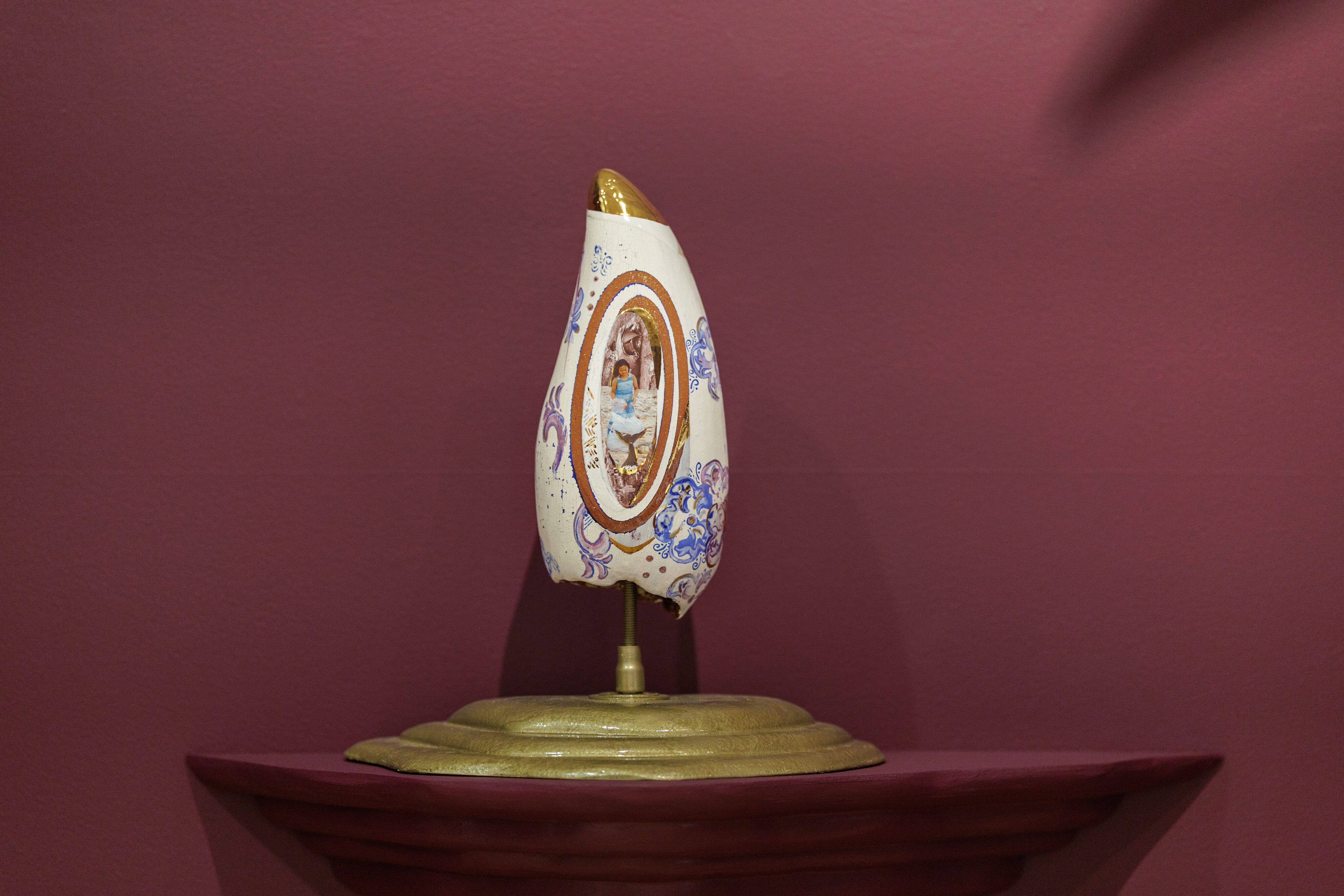A sculptural whale tooth displayed against a pink wall. The object has an elongated, oval shape with a gold-painted top and intricate blue and white patterns on its surface. In the center, there is an oval illustration of a person in a blue dress, framed with gold and brown. The object is mounted on a stand, showcasing its detailed and artistic design.