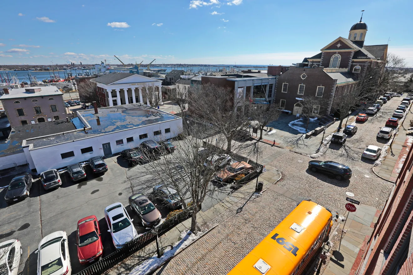Aerial view of cobblestone streets, New Bedford Whaling Museum, historic buildings, parked cars, and harbor.