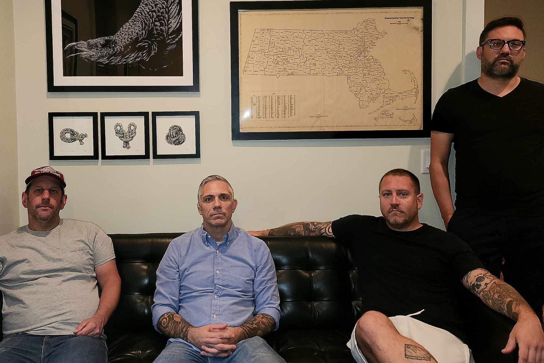 Wire Lines Four men sitting on a couch with framed art and a map on the wall behind them.