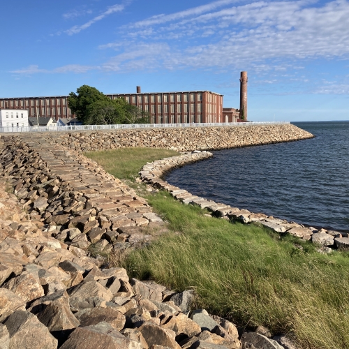 Image of New Bedford Hurricane Barrier with rock walls and grass between a large brick building and water.