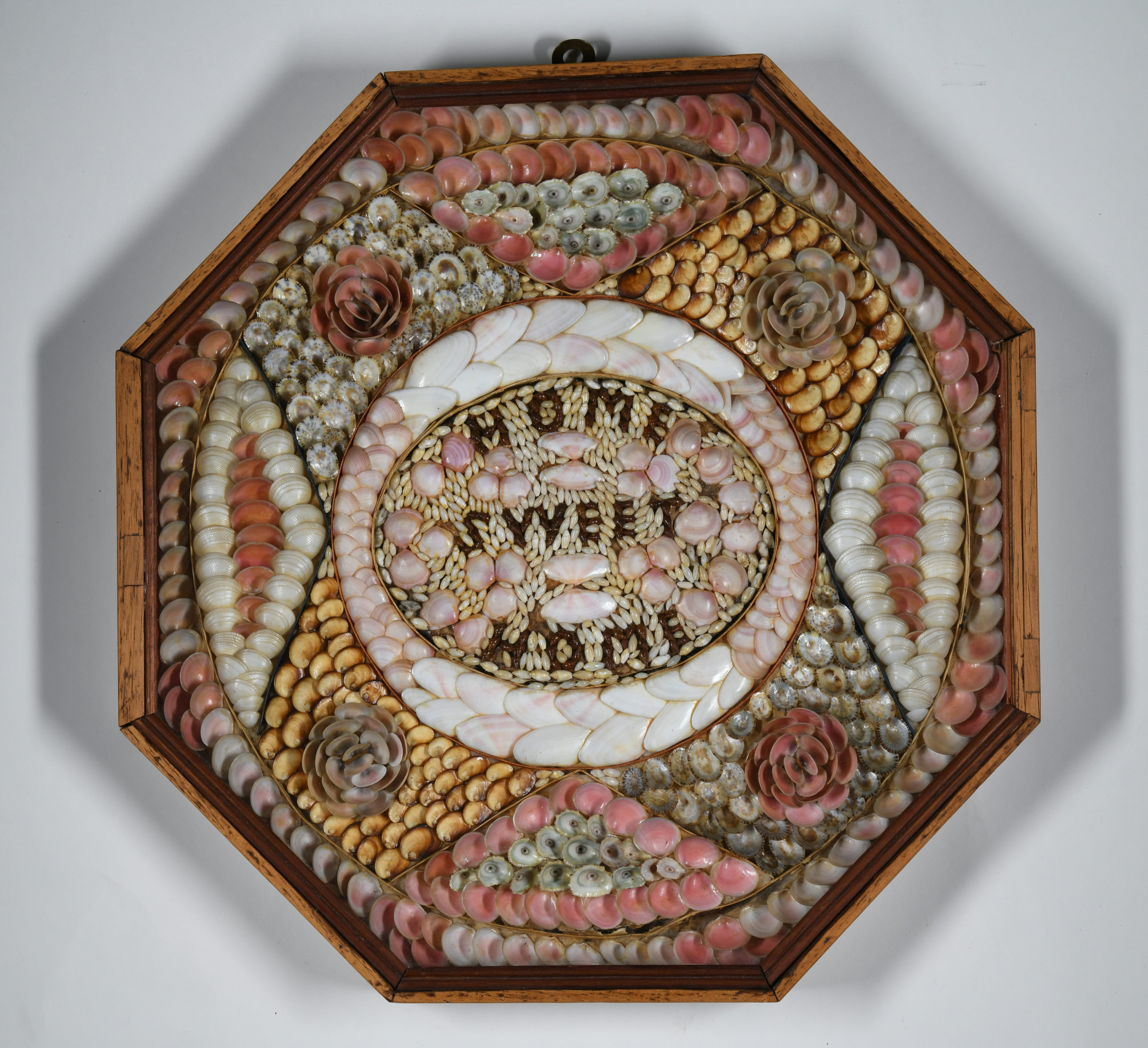 An octagonal shadow box containing a Victorian sailor's valentine with the words "HOME SWEET HOME" spelled out in small seashells at the center. The surrounding design features intricate, concentric patterns of pink, white, and tan seashells arranged in floral and geometric shapes.