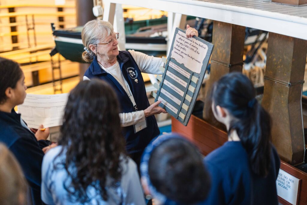 A museum volunteer at the New Bedford Whaling Museum holds an educational board for a group of students, illustrating the costs deducted from a whaler's earnings after a voyage.