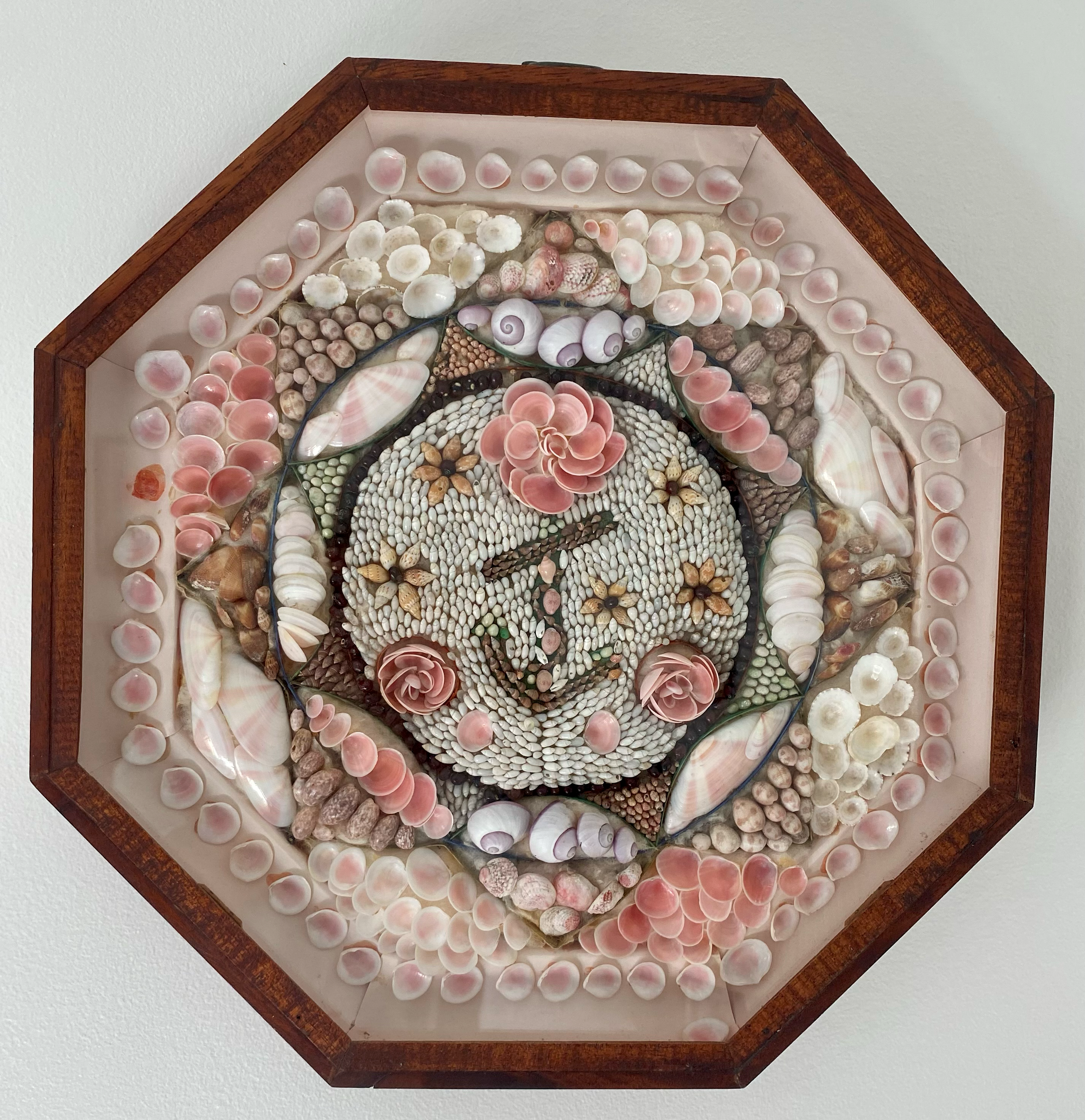 An octagonal shadow box containing a Victorian sailor's valentine, featuring intricate patterns of pink and white seashells arranged on a light background. In the center, shells are mosaic-tiled to form an anchor and floral motifs.